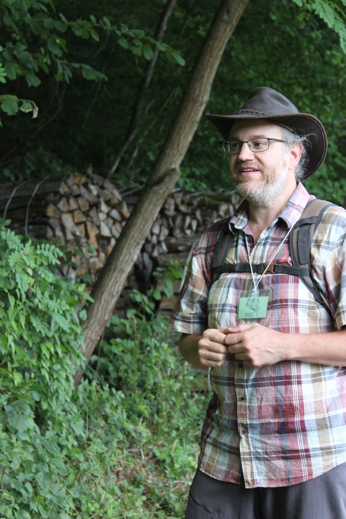 Photo de Nicolas à l'orée de la forêt.  Il porte une chemise à carreaux, un chapeau, un badge et un sac-à-dos. Il se tient debout devant des arbres et des stères de bois.  Il parle et regarde devant lui, vers la gauche de la photo.