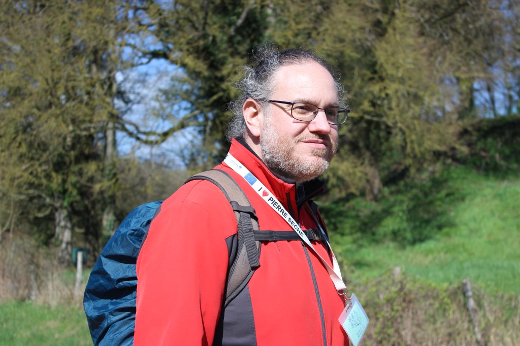Photo de Nicolas de trois-quarts.  Il regarde devant lui, vers la droite, avec un léger sourire. Il porte un blouson rouge et un tour de cou I love pierre sèche au bout duquel on devine son badge, ainsi qu'un sac-à-dos recouvert d'une protection imperméable. Il se tient debout ; derrière lui, on devine une prairie avec une palissade, puis des arbres aux branches encore nues.